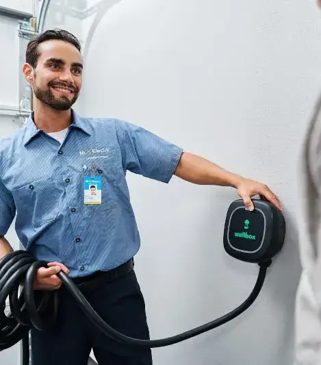 A Mr. Electric Electrician Holds a Coiled Cord Attached to an EV Charger on the Wall and Touches it to Show How to Use It.