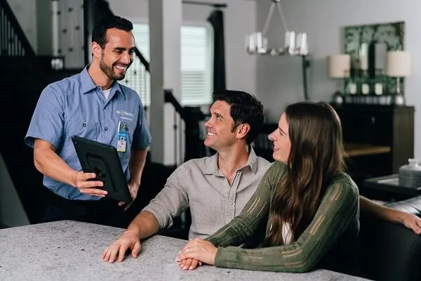 A Smiling Mr. Electric Electrician Shows a Tablet to a Seated Man and Woman at a Counter Next to Him