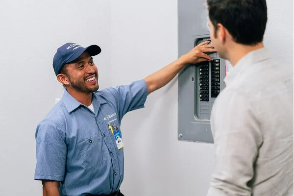 A smiling Mr. Electric electrician points at an open electrical panel while talking to a man.