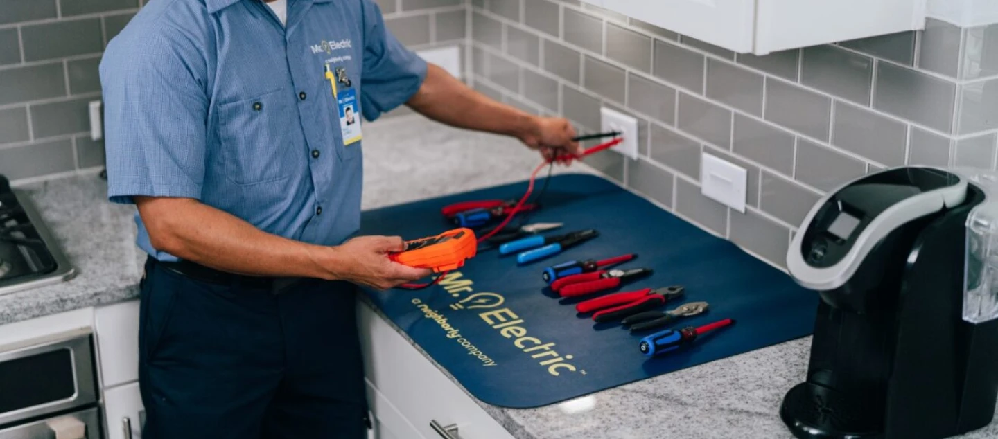 A Mr. Electric electrician using an electrical outlet tester in a kitchen during a home wiring upgrade appointment