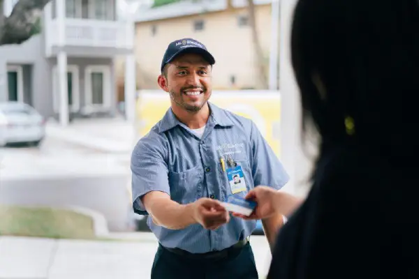 A Woman in Shadow Pictured from Behind Take a Business Card from a Smiling Mr. Electric Electrician Standing Outside Her Door.