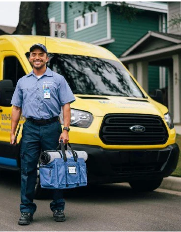 A smiling Mr. Electric electrician stands in front of a Mr. Electric van holding a bag with a rolled door mat on top of it.