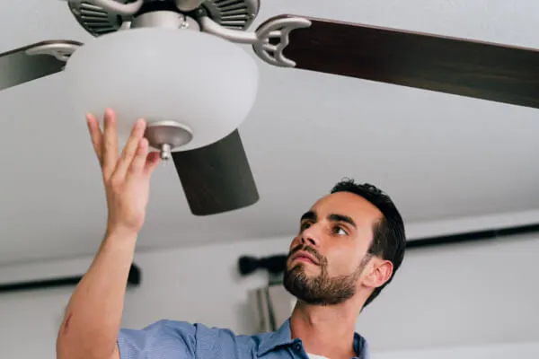 A Mr. Electric electrician reaches up to place his hand beneath the light fixture attached to a ceiling fan.