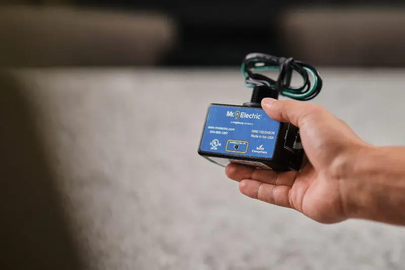 A Hand Holds a Black House Surge Protector with Coiled Cords on the Top and a Blue Label with a Mr. Electric Logo on It