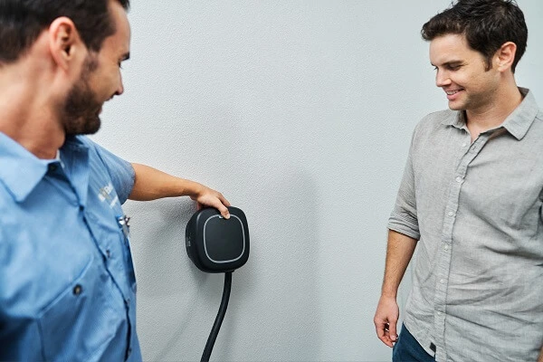 A Mr. Electric electrician holds a coiled cord attached to an EV charger on the wall and shows another man how to use it.