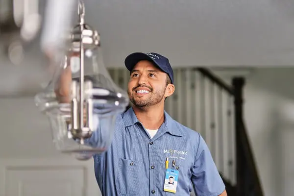  A smiling Mr. Electric electrician reaches up to a pendant light fixture hanging from a ceiling. 