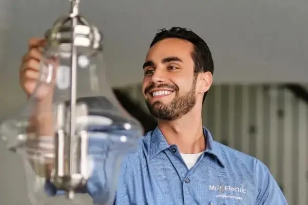 A smiling Mr. Electric electrician reaches out to a pendant light fixture suspended from a ceiling.