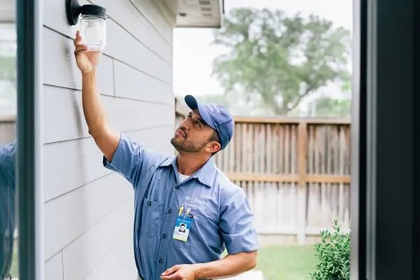 A Mr. Electric electrician reaches up to grasp the glass cover on an exterior light fixture. 