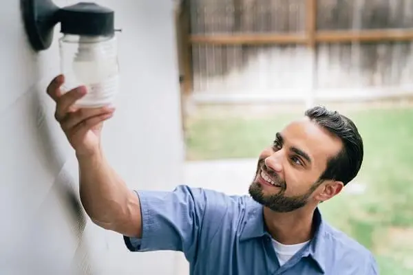 A Smiling Mr. Electric Service Professional Reaches Up to Grasp the Glass Cover on an Exterior Light Fixture.