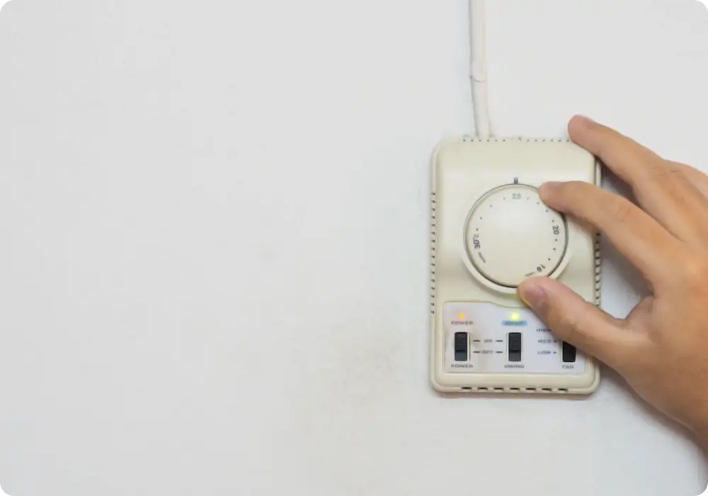 A right hand is shown adjusting the dial on a rectangular, off-white thermostat mounted on a plain white wall. The thermostat has a circular dial with temperature markings and several small indicator lights and buttons below.