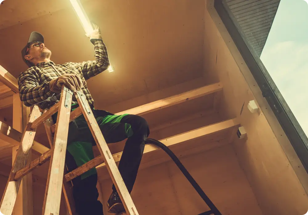 Low angle view of a man wearing a plaid long-sleeved shirt, safety glasses, a dark baseball cap, and green and black work pants, standing on a wooden ladder and installing a long fluorescent light fixture into the wooden framework of a building under construction.