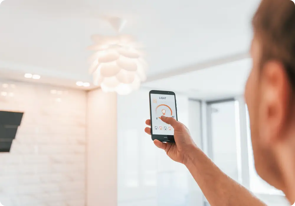 A hand holds a black smartphone displaying a light control interface, pointed toward a modern white flower-shaped ceiling light in a room with white brick walls.