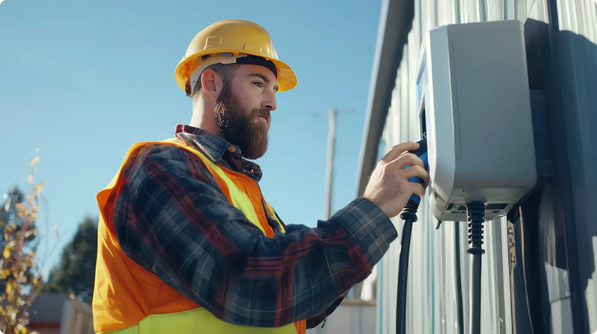 A bearded electrician wearing a yellow hard hat, a red and black plaid shirt, and a bright yellow safety vest is connecting a charging cable to a white electric vehicle charger mounted on a gray building exterior. The sky is clear and blue. Part of a residential area with houses and trees is visible in the blurred background.