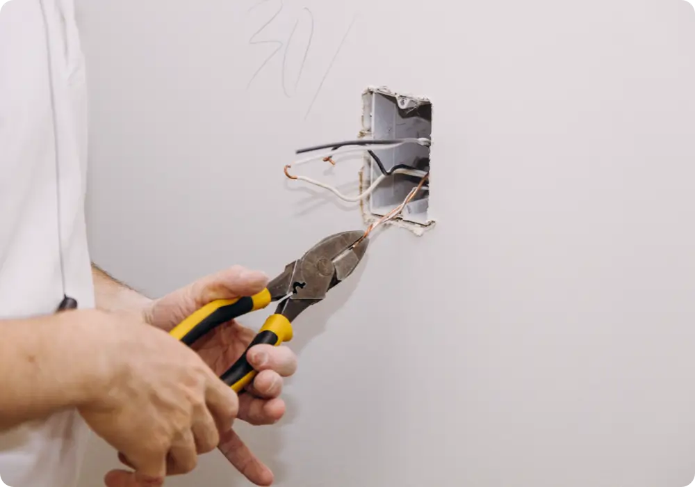 A person wearing a white shirt is using yellow-handled wire cutters to trim electrical wires inside a wall socket box in a white wall.