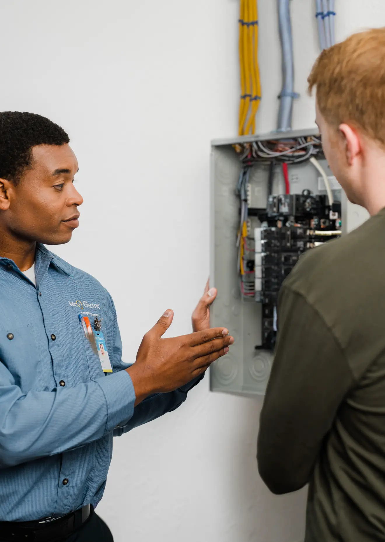 Electrician in blue shirt explaining the components of an open electrical panel to a customer in a home with visible wiring and circuit breakers.