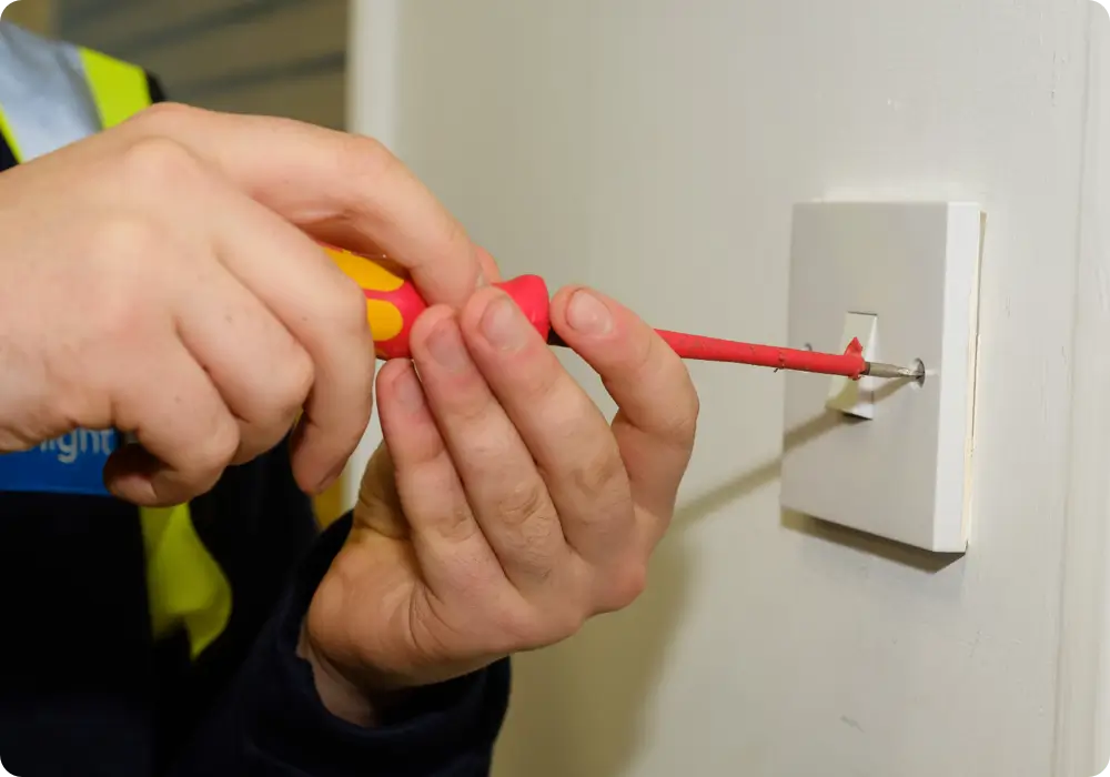 Close-up of a person's hands, wearing a dark long-sleeved shirt and a neon yellow safety vest detail, using a red and yellow Phillips head screwdriver to secure a white light switch to a light beige wall.