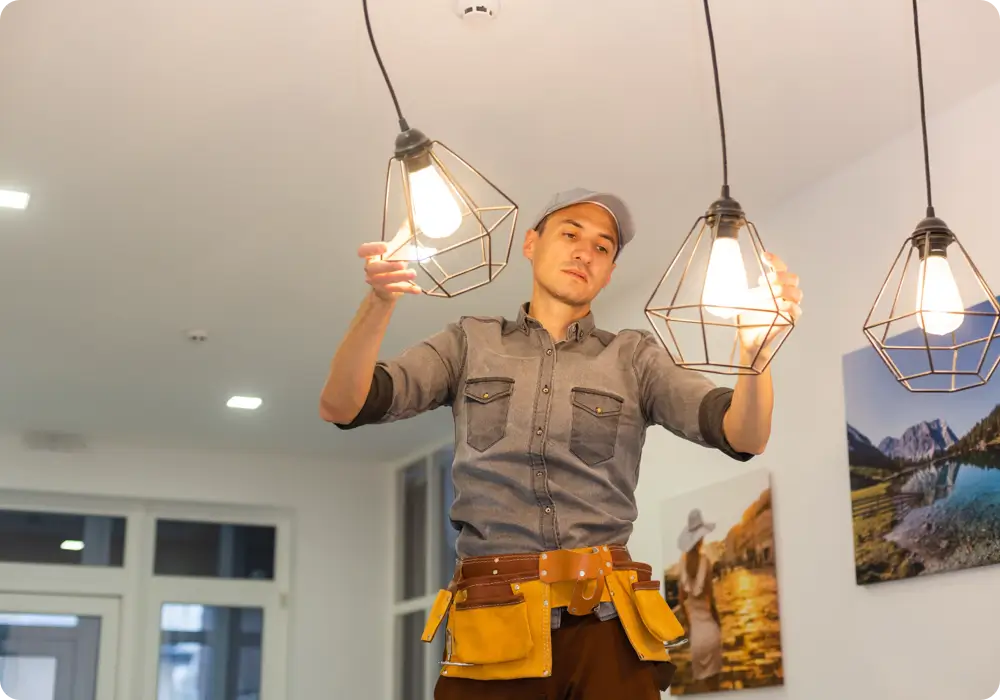 An electrician wearing a grey cap, a long-sleeved grey work shirt, and brown trousers with a yellow and brown tool belt is installing three modern geometric pendant lights with exposed bulbs in a room.