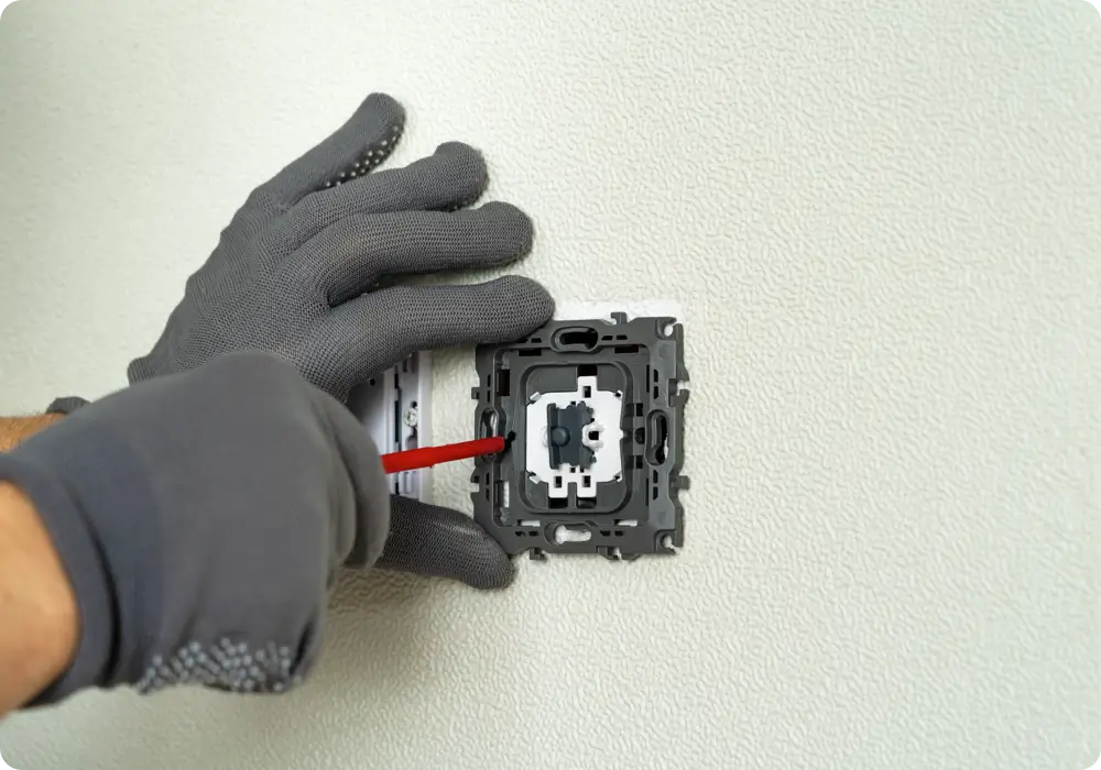 Close-up of two hands wearing grey work gloves with black dots, installing the internal mechanism of a white light switch onto a textured off-white wall using a red handled screwdriver.