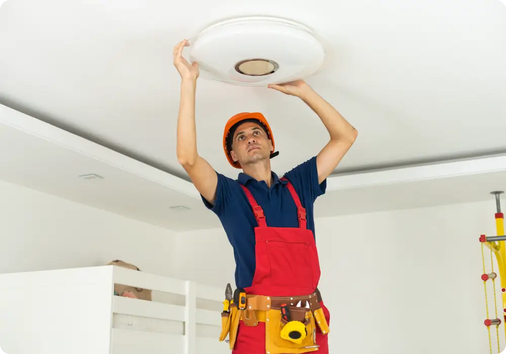 A handyman wearing an orange hard hat, a blue polo shirt, and red overalls with a tool belt is installing a round, frosted white LED ceiling light fixture on a white ceiling in a room with white walls.