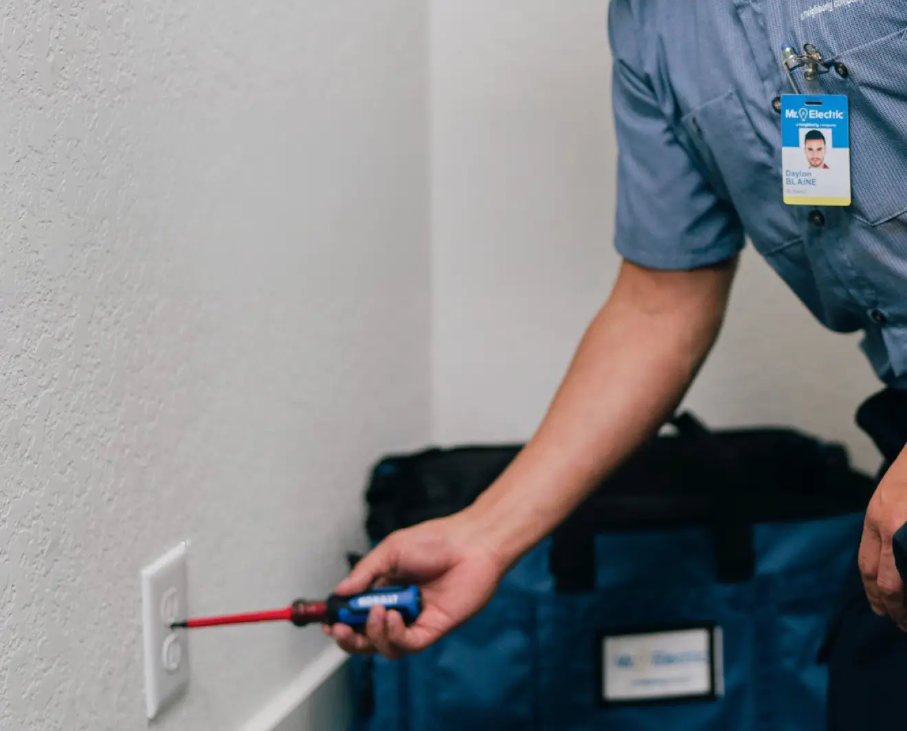 Handyman in uniform using a screwdriver to install or repair a commercial-grade electrical outlet on a white wall, with a blue tool bag in the background.