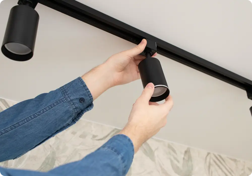 Two hands wearing a long-sleeved blue denim shirt are shown installing a black track lighting fixture onto a white ceiling. Another similar light fixture is already attached to the left.