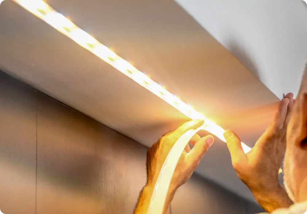 Close-up of two hands with short fingernails pressing and adhering a bright warm white LED strip light underneath a dark brown or black cabinet or shelf. The light illuminates the surrounding area.