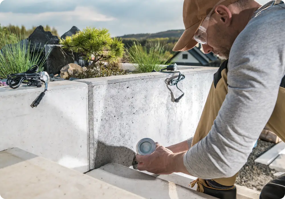 A man wearing safety glasses and a brown baseball cap is installing a small, round outdoor pathway light into a concrete retaining wall in a garden setting. He is wearing a grey long-sleeved shirt and tan work pants and kneeling on concrete steps.