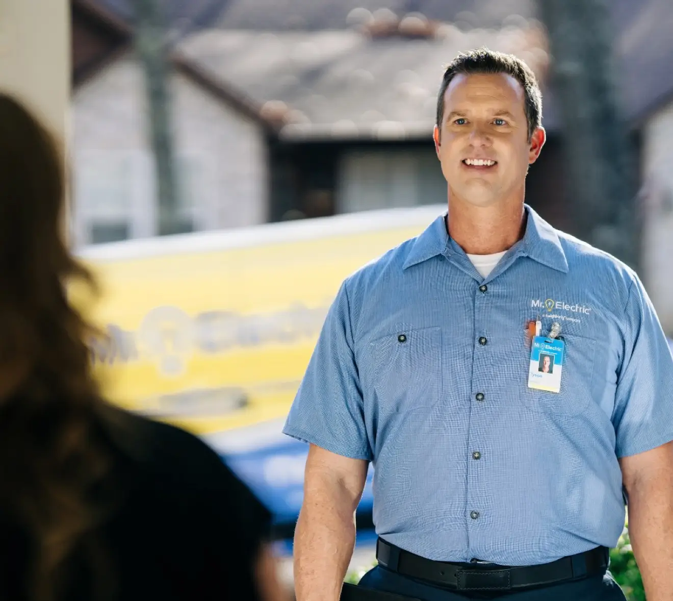 Professional electrician in blue Mr. Electric uniform smiling and greeting a customer outside a home, with a branded service van in the background.