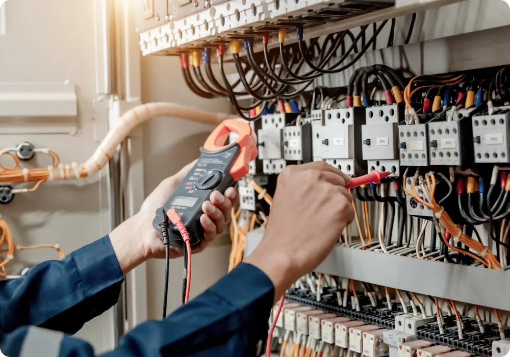 A technician wearing a dark blue long-sleeved shirt is using a digital multimeter with red and black probes to test the circuits inside a large electrical panel with numerous wires and components.