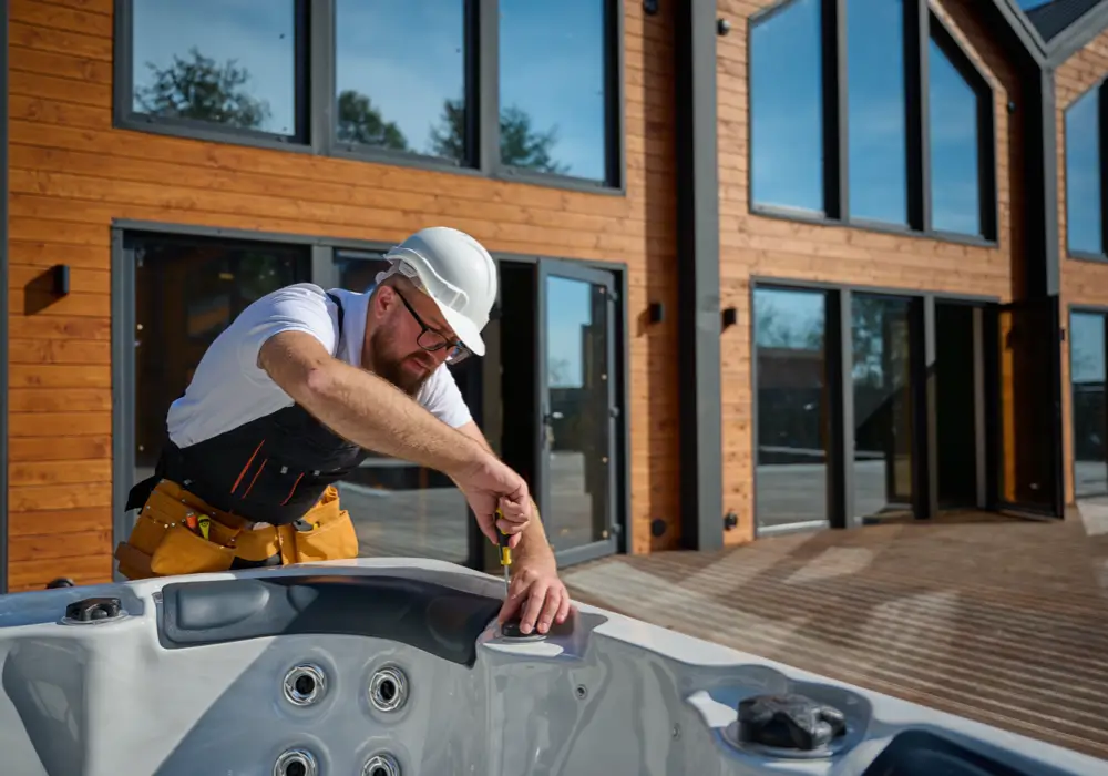 Construction worker wearing a white hard hat, glasses, white T-shirt, and yellow tool belt, using tools to install a gray hot tub outdoors, in front of a modern wooden house with large windows and a wooden deck.