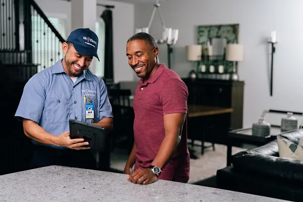 A Smiling Mr. Electric Electrician Looks Down and Points at a Tablet He’s Holding While a Smiling Man Beside Him Looks at It.