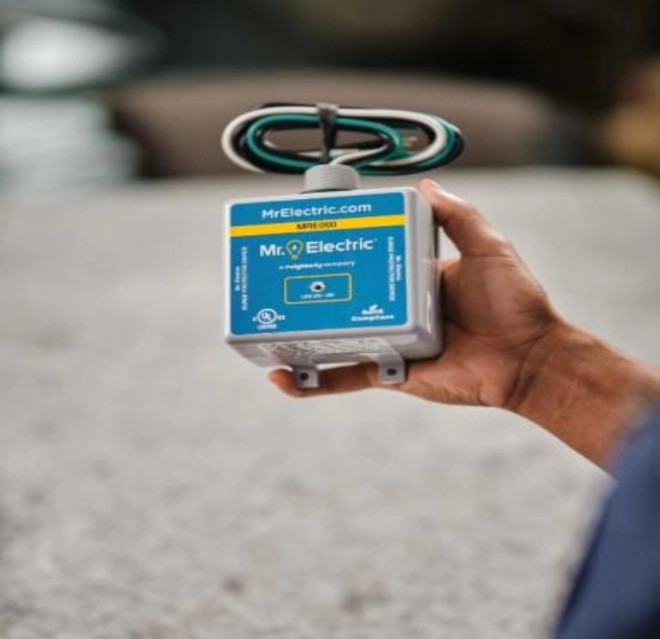 A Hand Holds a Black House Surge Protector with Coiled Cords on the Top and a Blue Label with a Mr. Electric Logo on It.