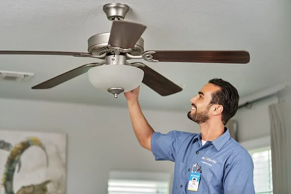 A smiling Mr. Electric Electrician reaches up to the top of a light fixture attached to a ceiling fan