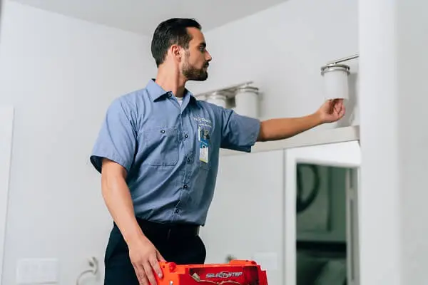 A Mr. Electric electrician on a ladder adjusts the bulb in a light fixture above a bathroom mirror. 
