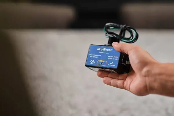 A hand holds a black house surge protector with coiled cords on the top and a blue label with a Mr. Electric logo on it  