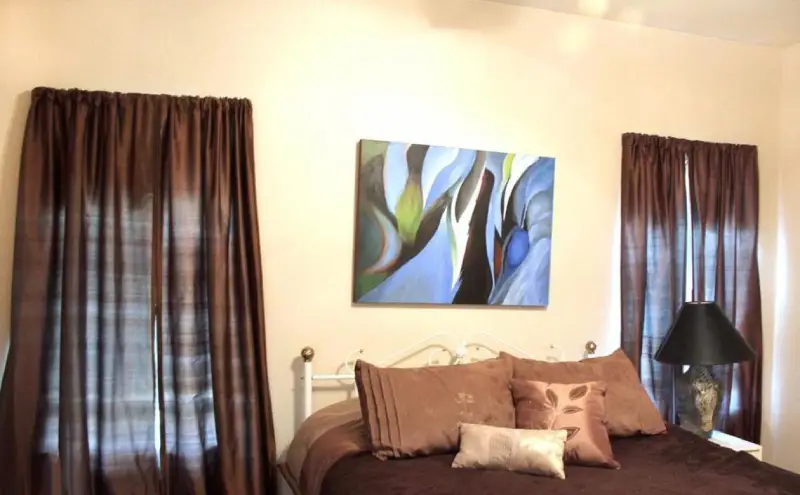 Photograph of a bedroom in a house with a bed in between two windows.  Both windows have brown curtains.