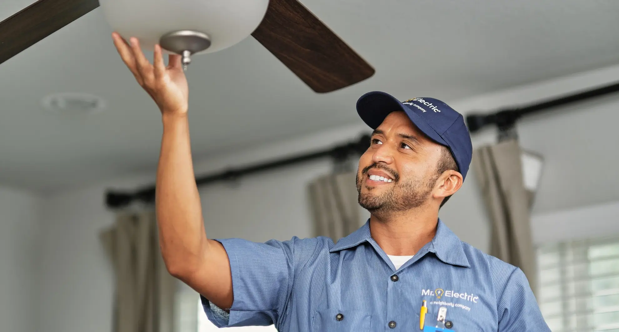 A smiling electrician in a blue shirt adjusts ceiling fan light inside a home.