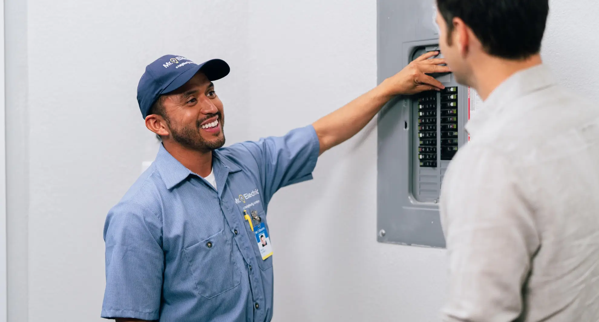 A smiling electrician in a blue uniform explains the breaker panel to a man in a light shirt indoors.